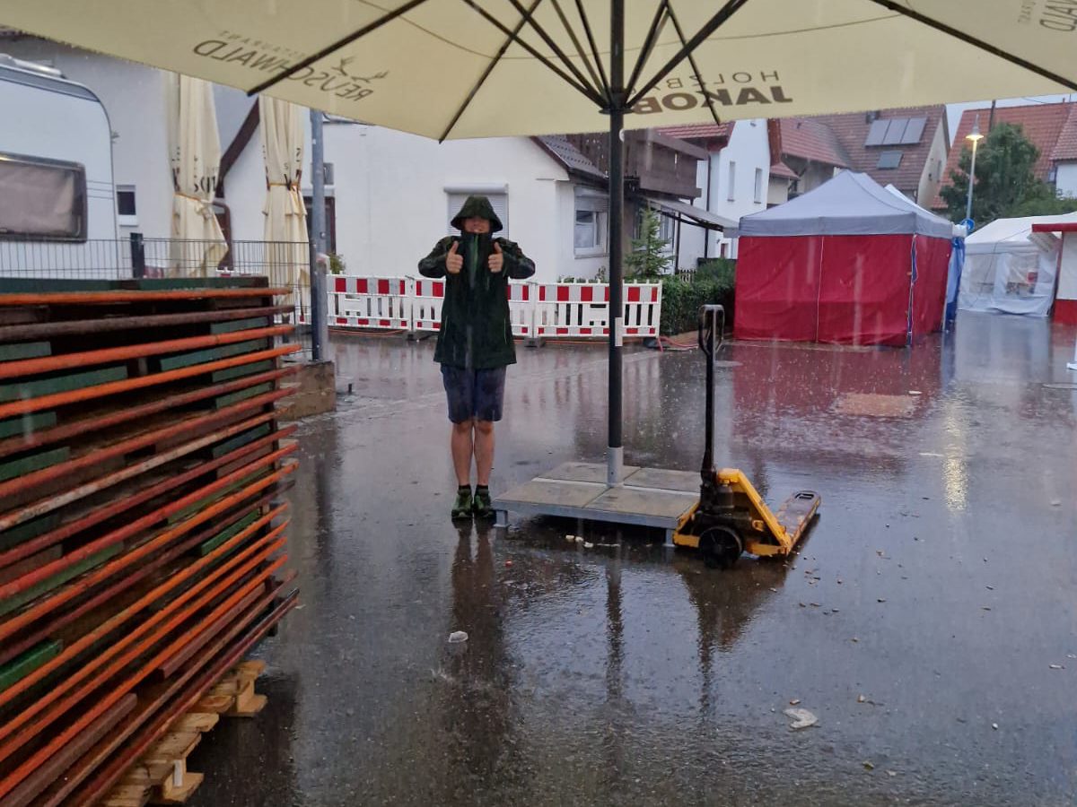 Im strömenden Regen hebt ein Helfer in Regenjacke unter einem Sonnenschirm beide Daumen, ringsum gestapelte Biertische