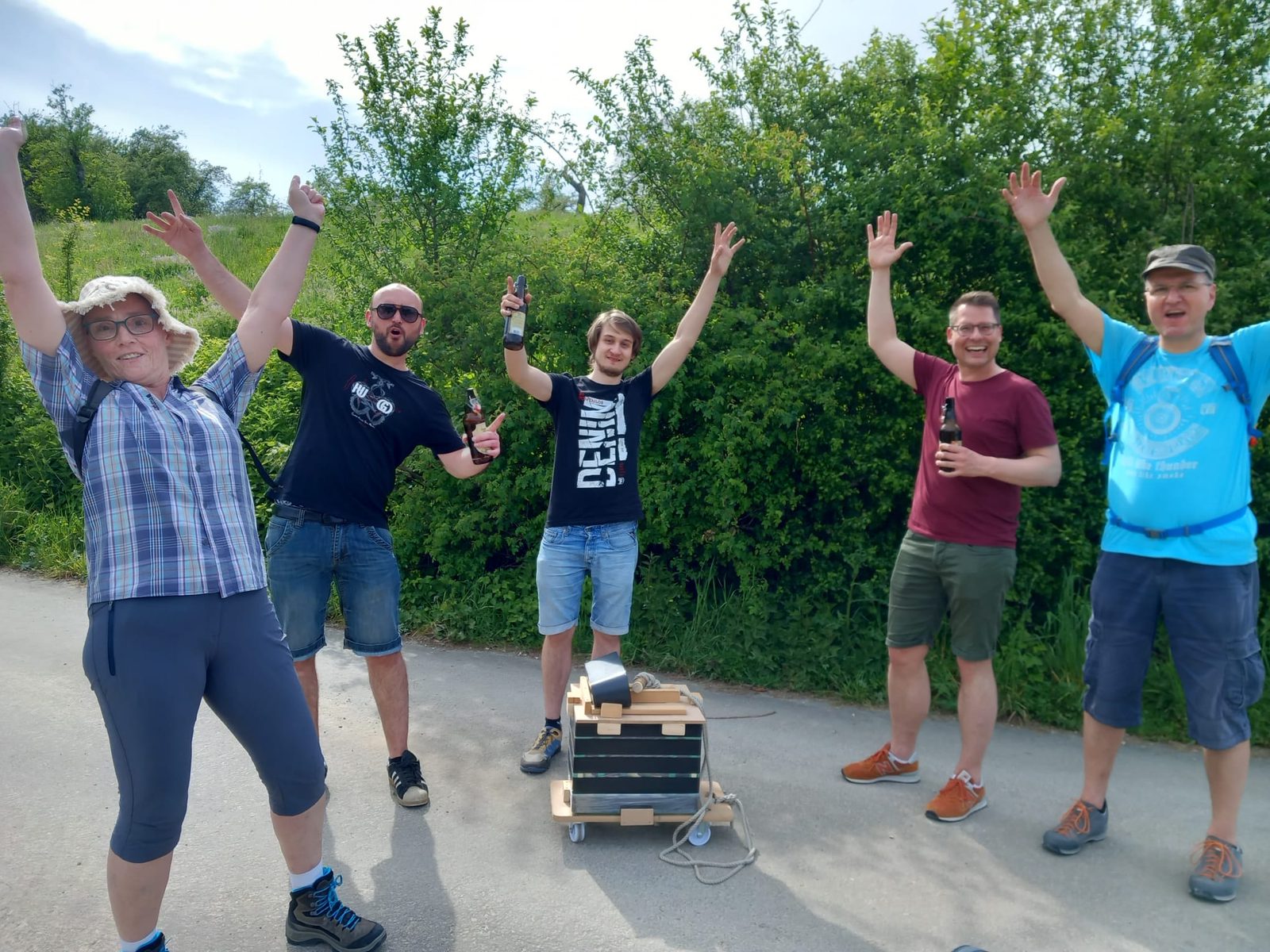 Fünf Wandernde stoßen vor einer Hecke an, in der Mitte der Bollerwagen mit Lautsprecher